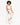 A model with curly hair sits on a stool, wearing a draped white top and matching jogger pants with white sneakers.