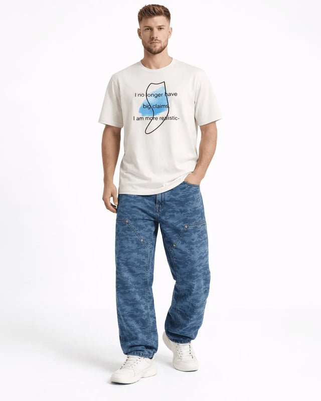 A man stands casually wearing a graphic t-shirt and loose-fitting camo pants, paired with white sneakers.