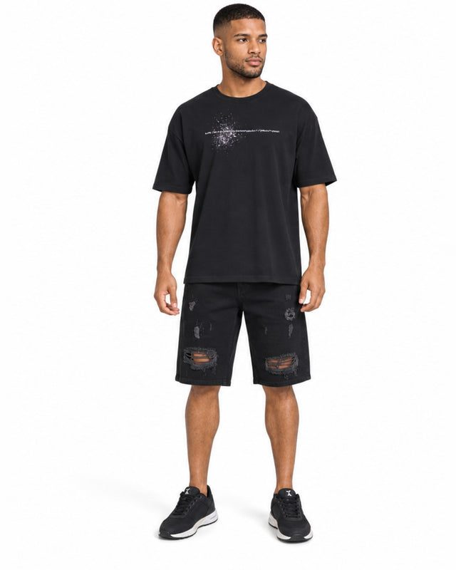 A man stands wearing a black graphic t-shirt and distressed black shorts, paired with black sneakers.