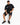 A man sits on a white cube wearing a black graphic t-shirt, distressed black shorts, and black sneakers.