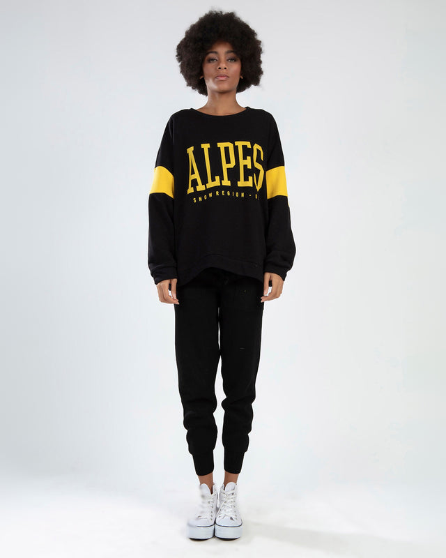 Model in black ALPES streetwear crop top with yellow graphics and stripe sleeves, paired with matching ribbed joggers.