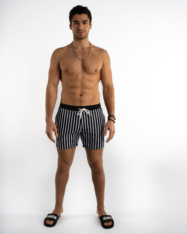 Man in Niko black and white striped swim trunks with white drawstring, Carbon brand slides, and poolside summer style.