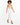 A model with curly hair wears a fitted beige top and white tailored pants, paired with black high-heeled sandals.