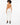 A woman with curly hair wears a tan bodysuit with a stylish back design, paired with white pants and black heels.