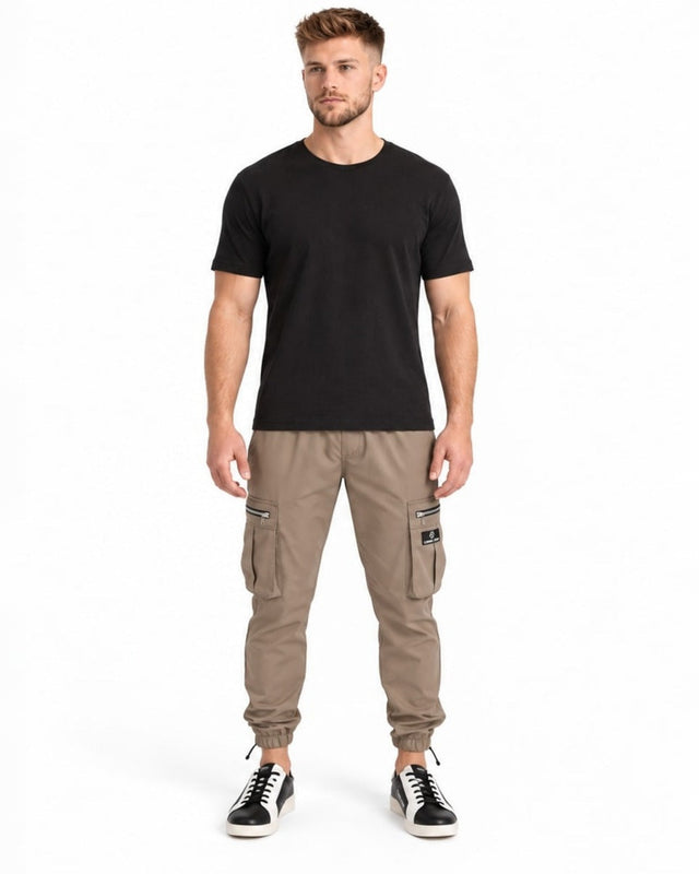 A man stands wearing a black t-shirt and beige cargo pants with zippered pockets and elastic cuffs.