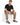 A man sits on a white cube wearing casual khaki pants and black sneakers, paired with a black t-shirt.