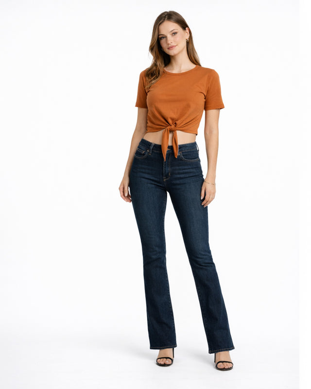 A woman stands confidently in a tied, rust-colored top and dark flared jeans, paired with black heels.