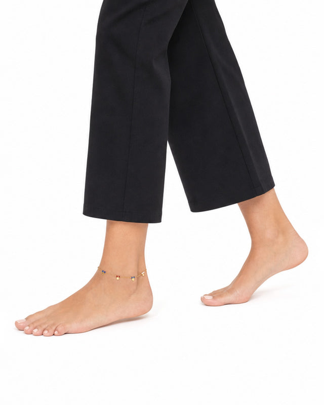 A bare foot with a delicate gold anklet featuring colorful charms, paired with black cropped pants.