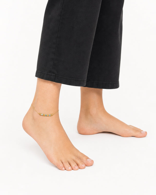 Delicate gold anklet with colorful beads, worn on a bare foot against a white background.