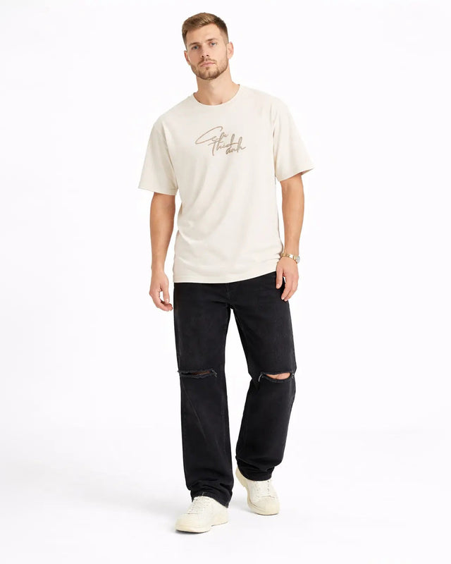 A man stands wearing a light beige t-shirt with a graphic design, paired with black ripped jeans and white sneakers.