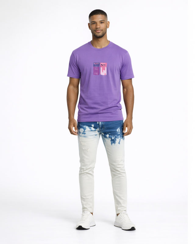 A man stands wearing a purple t-shirt and light blue jeans with a tie-dye pattern, paired with white sneakers.