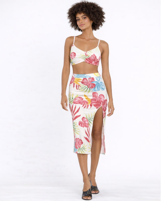 A model poses in a floral two-piece outfit featuring vibrant colors and a thigh-high slit, paired with black sandals.