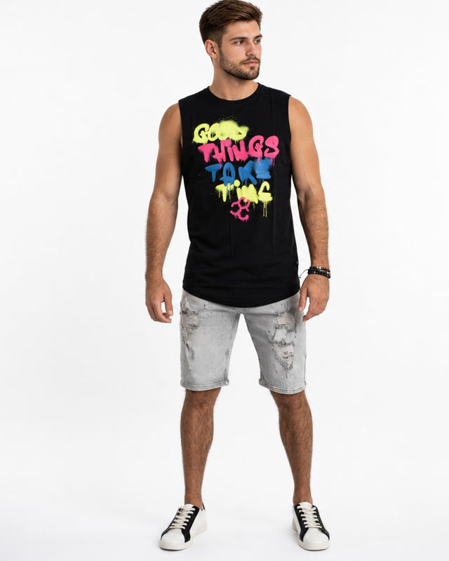 A man stands wearing a sleeveless black top with colorful graffiti-style text, paired with distressed gray shorts and...