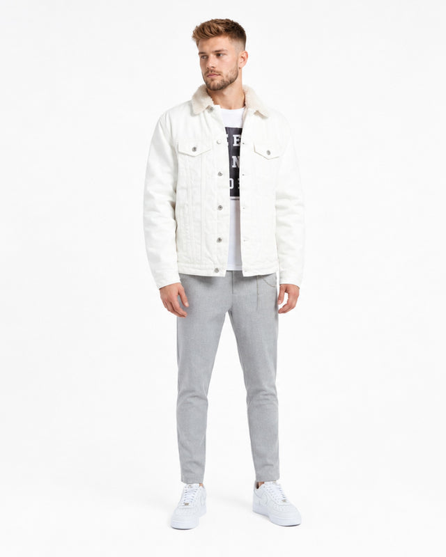 A man stands wearing a white denim jacket with a fluffy collar, paired with gray pants and white sneakers.