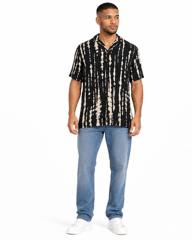 A man stands wearing a short-sleeve, black and beige patterned shirt paired with light blue jeans and white sneakers.