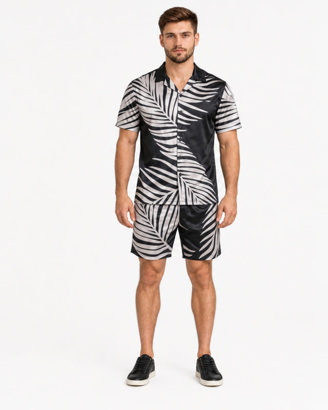 A man stands wearing a short-sleeve shirt and matching shorts with a black and white palm leaf pattern.