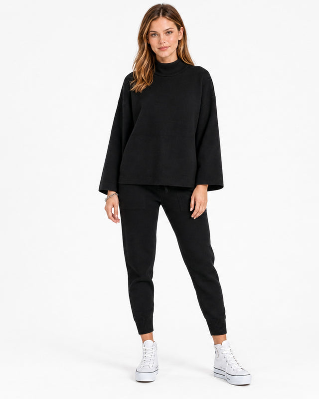 A woman stands confidently in a black, oversized sweatshirt paired with matching joggers and white sneakers.