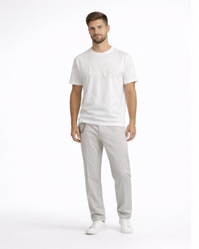 A man stands confidently in a white t-shirt with a textured graphic, paired with light gray pants and white sneakers.