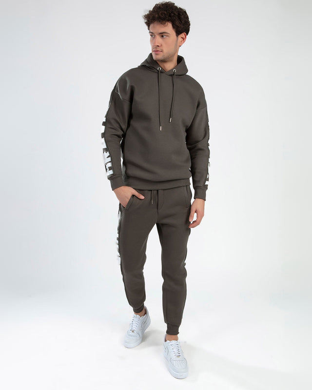 A model wearing a dark green hoodie and matching joggers, paired with white sneakers, stands against a plain background.
