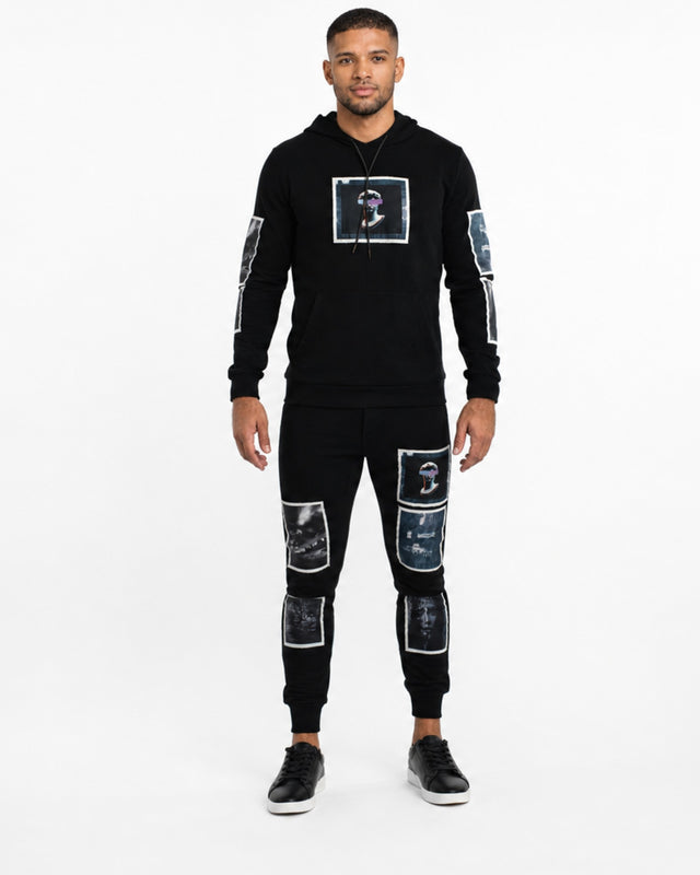 A man stands wearing black sweatpants with graphic patches and a matching hoodie, paired with black sneakers.
