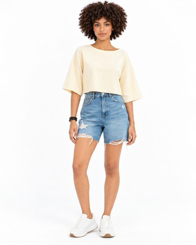 A model with curly hair wears a light-colored cropped top and distressed denim shorts, paired with white sneakers.