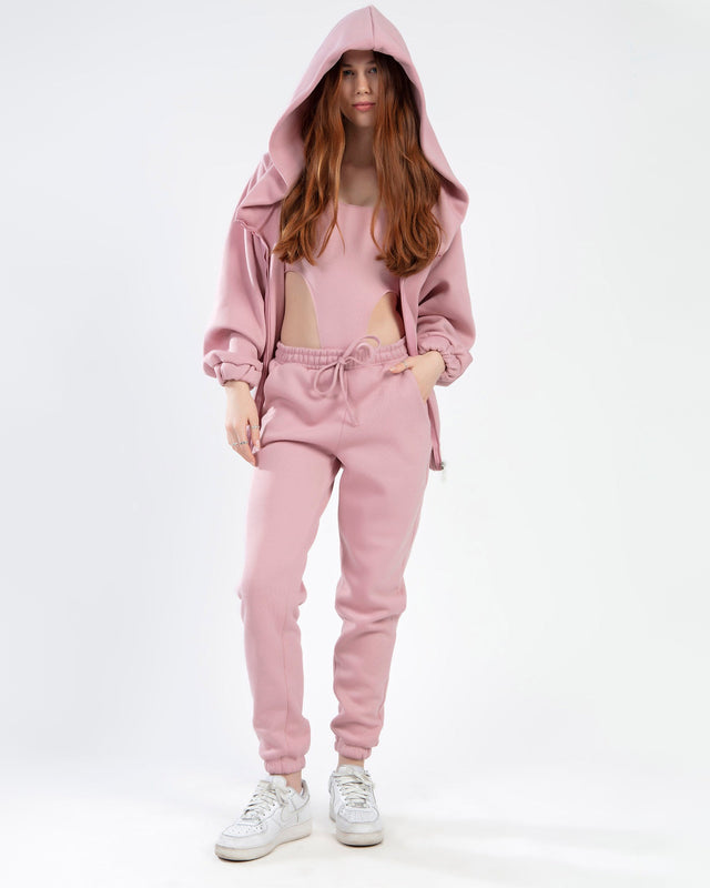 A model wearing a pink hoodie and matching joggers, paired with white sneakers, stands against a plain background.