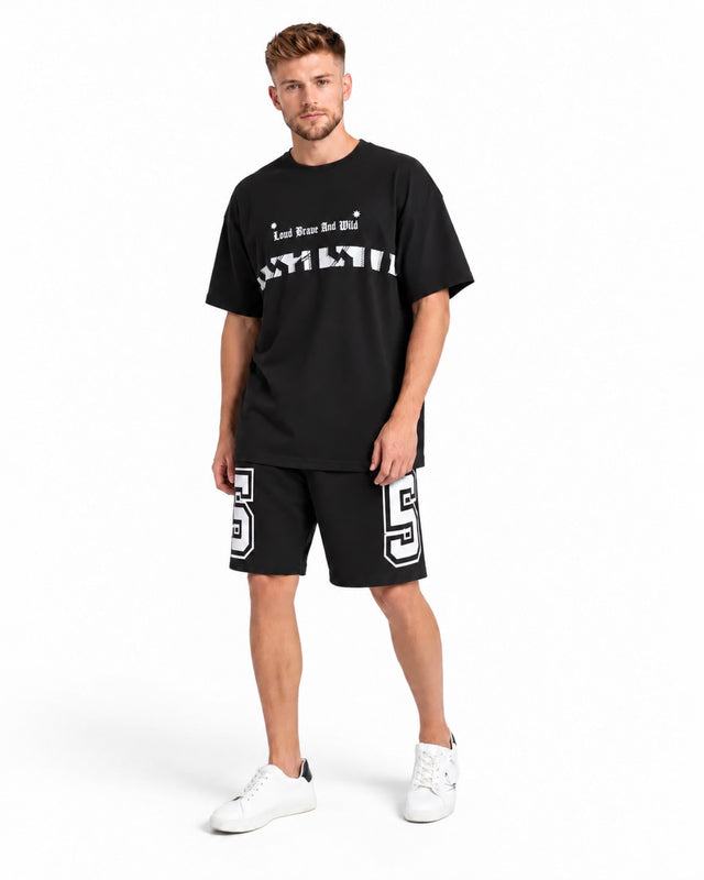 A man stands wearing a black oversized t-shirt with graphic text, paired with black shorts featuring a white number.