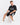 A man sits on a white cube wearing a black graphic t-shirt and black shorts with a white number design.
