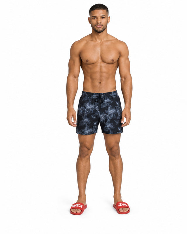 A shirtless man stands in swim trunks with a tropical leaf pattern, wearing red slide sandals.