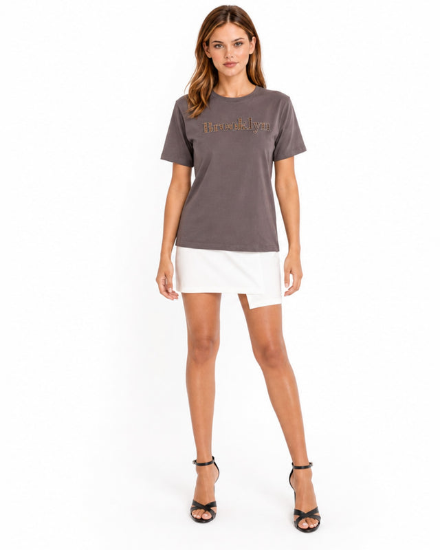 A woman stands confidently in a gray t-shirt with a stylish graphic, paired with a white skirt and black heels.
