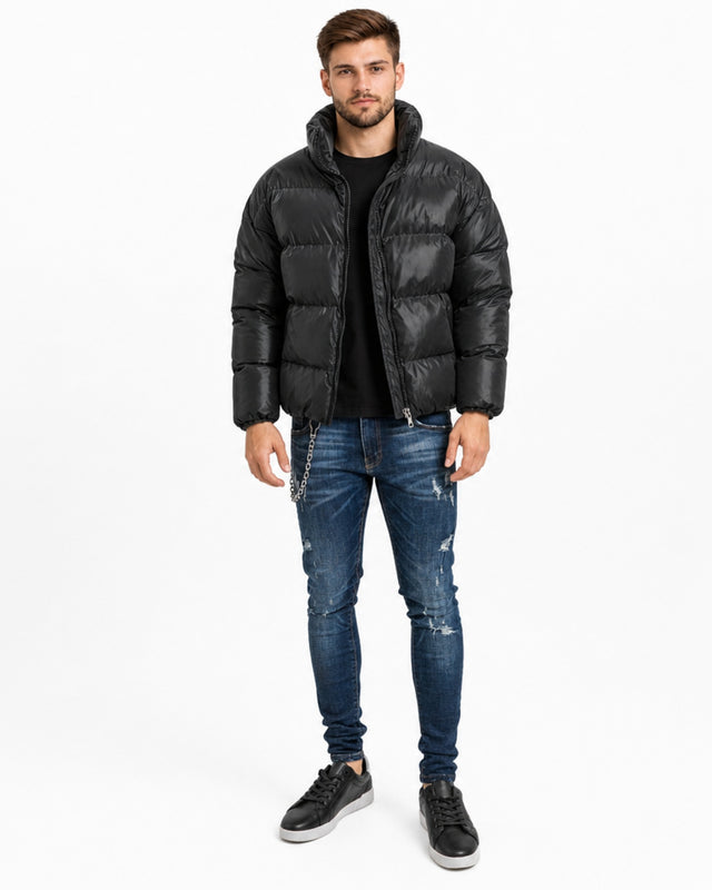 A man stands wearing a black puffer coat, black shirt, distressed jeans, and sneakers against a white background.