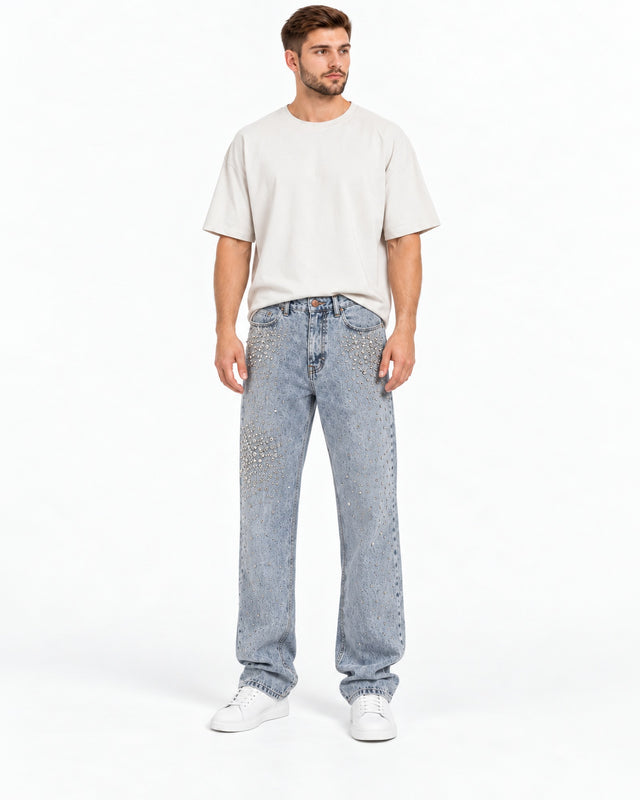 A man stands wearing a loose-fitting beige t-shirt and embellished light blue jeans.