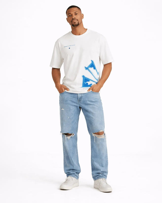 A man stands confidently wearing a white graphic t-shirt with blue designs and light blue distressed jeans.