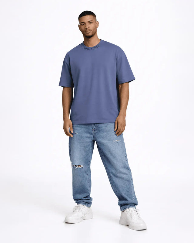 A man stands confidently wearing a blue oversized t-shirt and distressed light blue jeans with white sneakers.