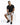 A man sits on a white pedestal wearing a black t-shirt, gray shorts, and black sneakers, showcasing a casual style.