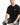 A man sits on a white pedestal wearing a black oversized t-shirt and distressed gray jeans, looking directly at the c...