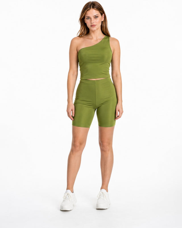 A model poses in a one-shoulder olive green top paired with matching bike shorts and white sneakers.
