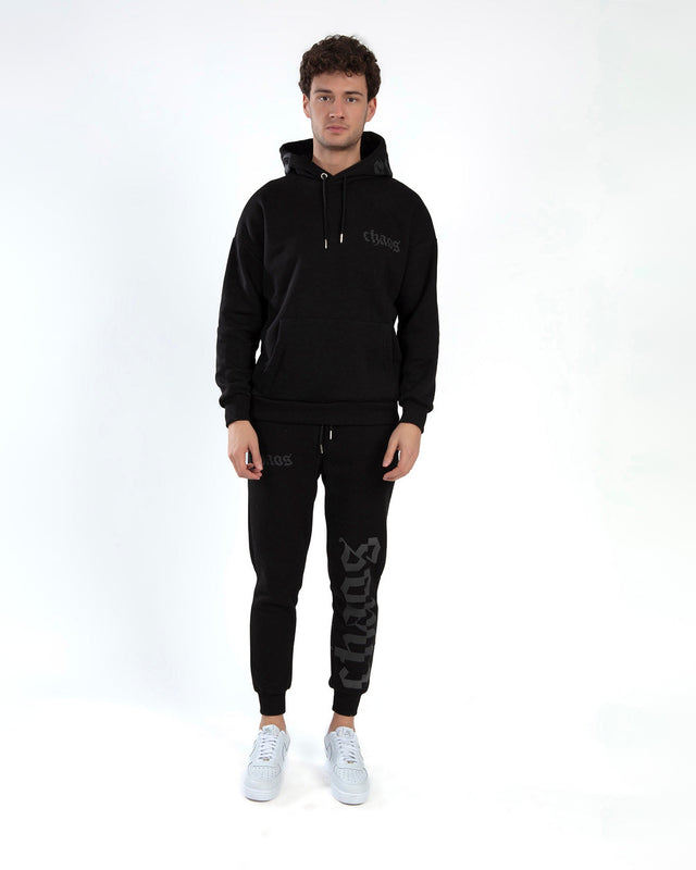 A young man stands wearing a black hoodie and matching joggers, paired with white sneakers.