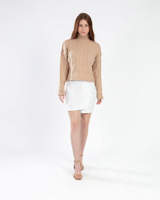 Model wearing Serena cable knit turtleneck sweater in tan with a white mini skirt and heels against a white background.