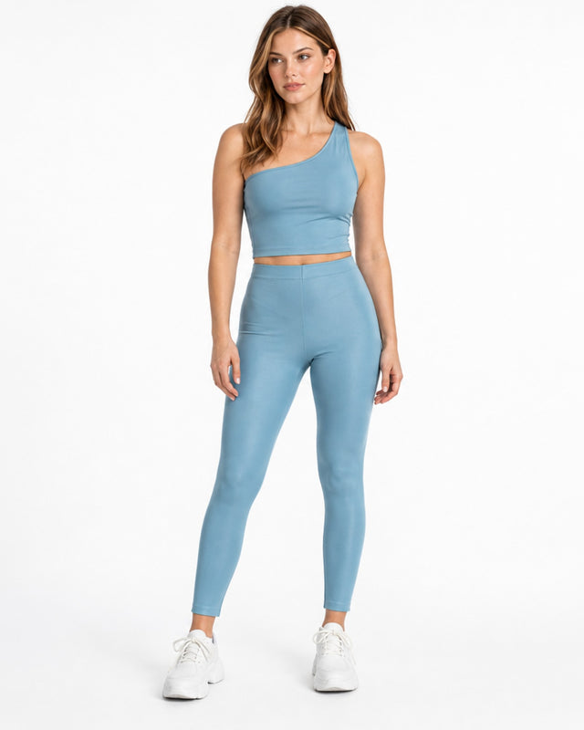 A woman stands confidently in a light blue one-shoulder crop top and matching leggings, paired with white sneakers.