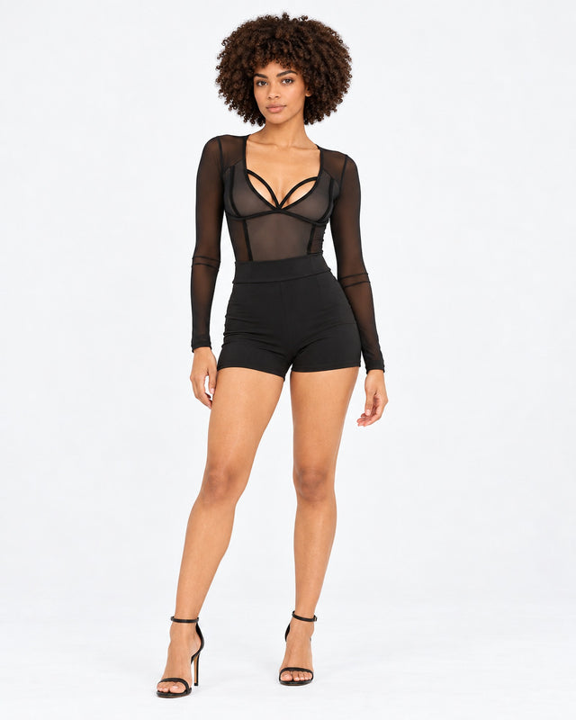 A model poses in a sheer long-sleeve top and high-waisted black shorts, showcasing a stylish, modern look.