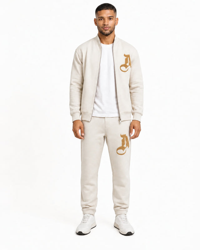 A man stands wearing a light beige zip-up jacket and matching sweatpants, featuring gold logos.