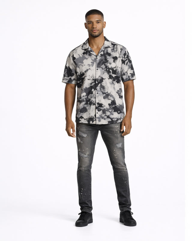 A man stands confidently in a short-sleeve, black and white patterned shirt paired with distressed jeans.