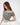 A model poses in a soft, off-the-shoulder, cropped top in a muted green shade, paired with white pants.