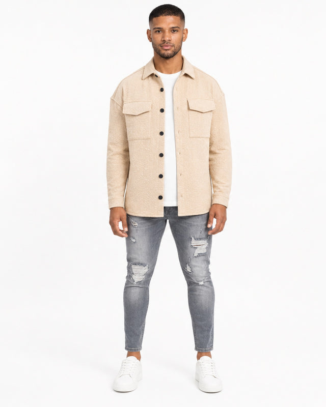 A man stands confidently in a light beige button-up top, paired with distressed gray jeans and white sneakers.