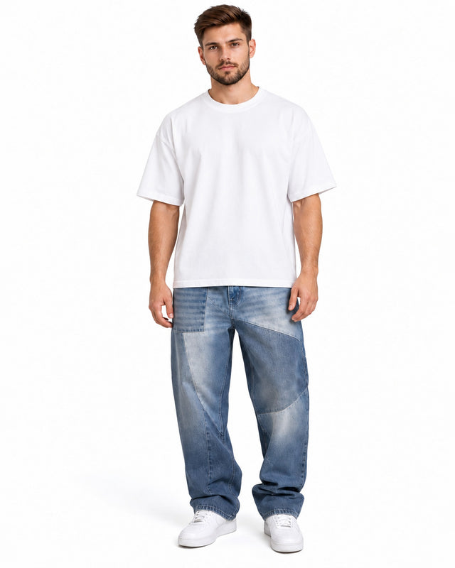 A man stands wearing a loose-fitting white t-shirt and light blue patchwork jeans, paired with white sneakers.