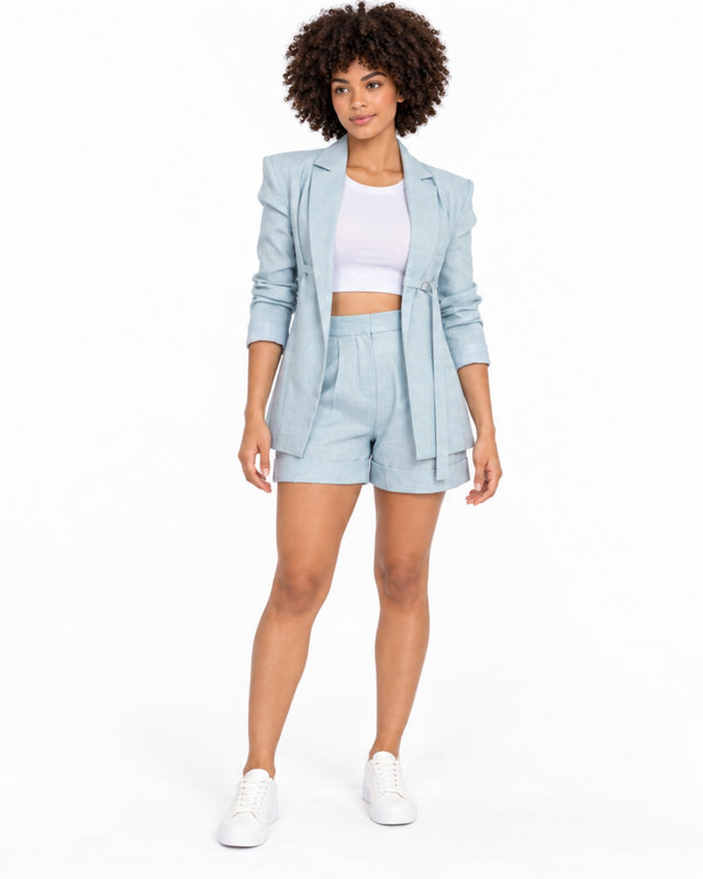 A woman with curly hair wears a light blue blazer and matching shorts, paired with a white crop top and sneakers.
