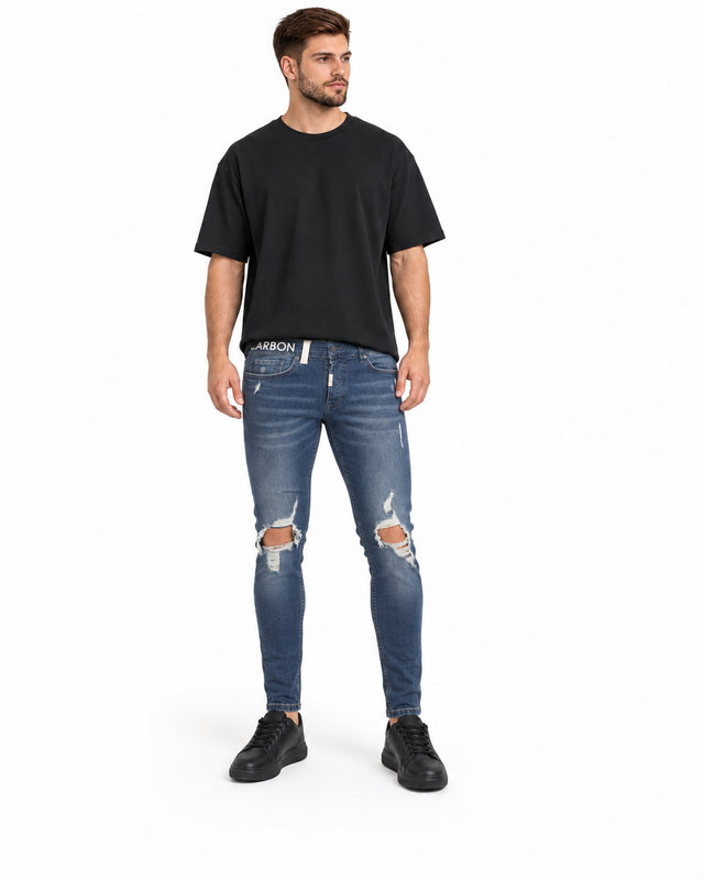 A man stands wearing a black oversized t-shirt, distressed blue jeans, and black sneakers.
