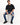 A man sits on a white pedestal wearing a loose black t-shirt, distressed blue jeans, and black sneakers.