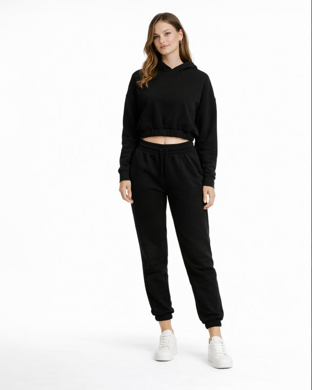 A woman stands confidently in a black cropped hoodie and matching joggers, paired with white sneakers.